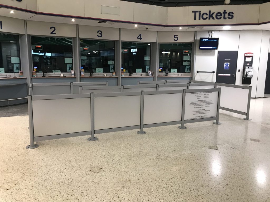 A Tensaguide® Plus Modular Barrier System with light grey infills and dark grey stanchions in front of a ticket office for crowd management.